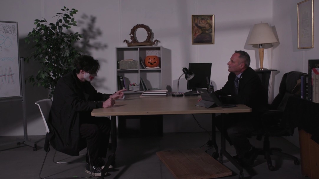 Two characters seated across a desk during an interrogation scene