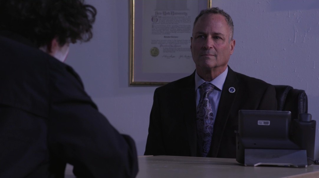 Man in a suit seated behind a desk during an interrogation scene